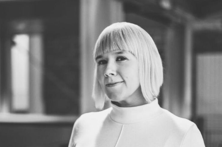 A headshot of a woman dressed in white with a short blonde bob and fringe, she smiles gently at the camera