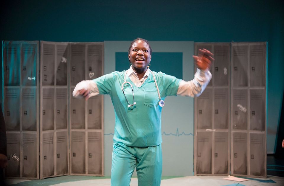 Performer in teal medical scrubs with a stethoscope gestures energetically on stage, standing in front of a backdrop of lockers in a hospital-themed set.