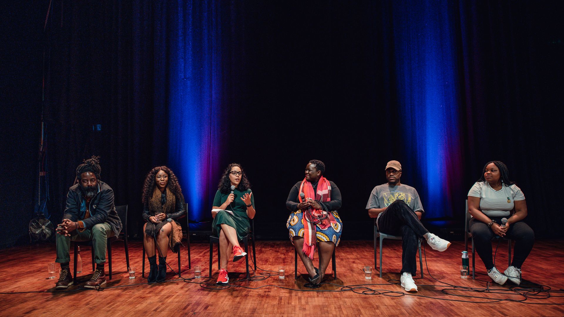 Six people sit on a panel on chairs in a row, the person in the middle holds a microphone