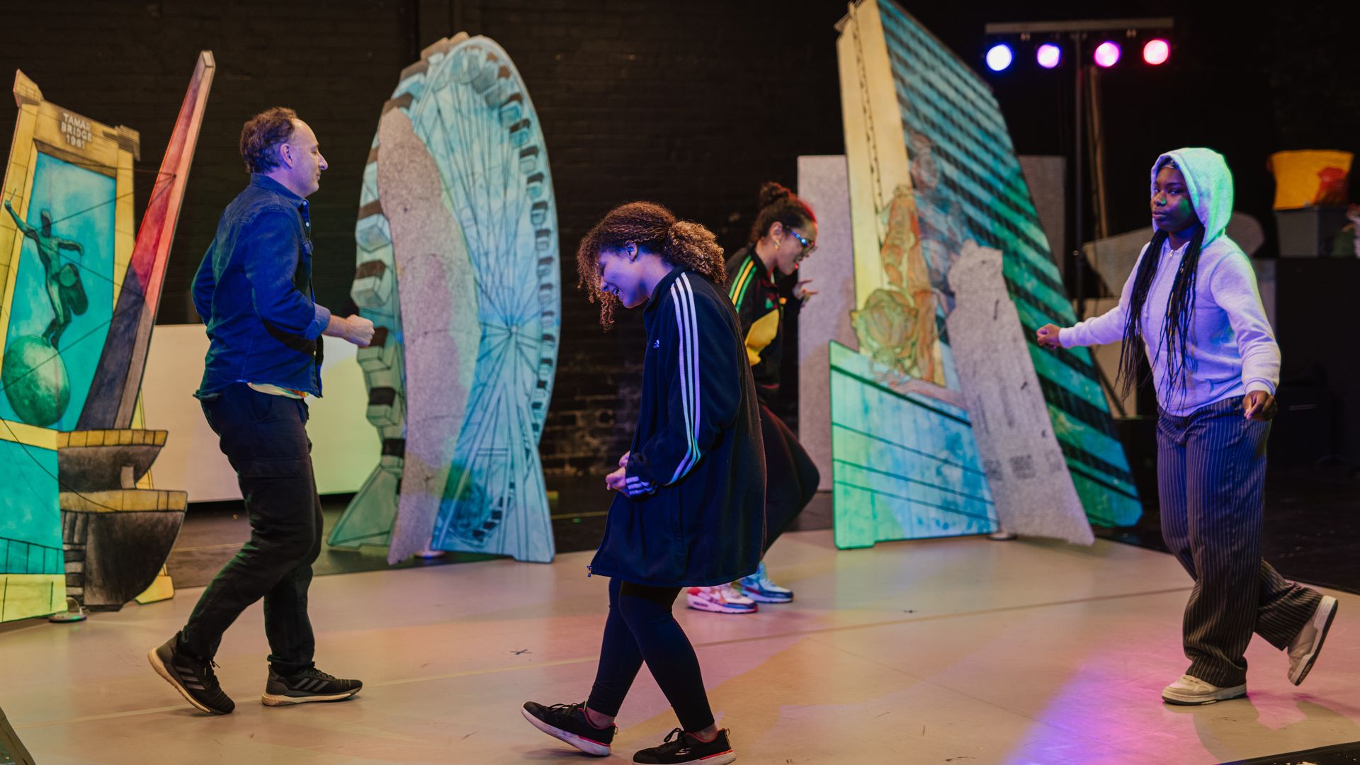 4 people dance in a circle on a stage. The stage includes props of buildings making it look like a city-scape