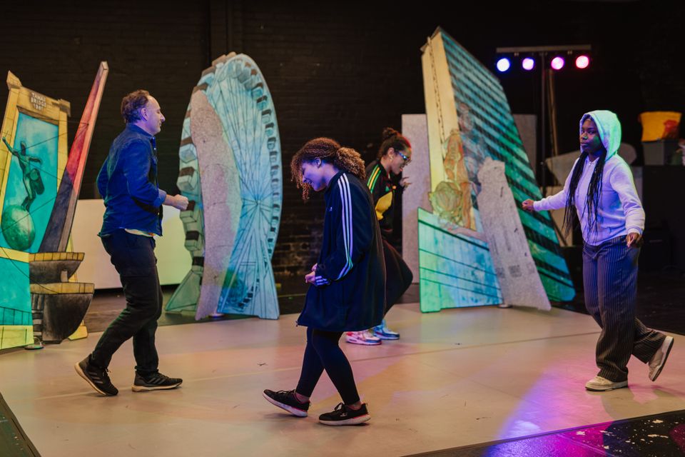 4 people dance in a circle on a stage. The stage includes props of buildings making it look like a city-scape