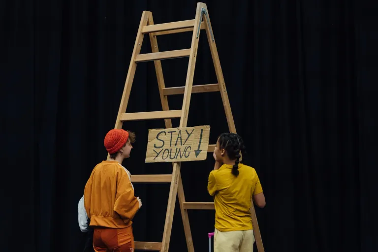 Two people are looking at a prop - a ladder with the sign 'Stay Young' stuck to it
