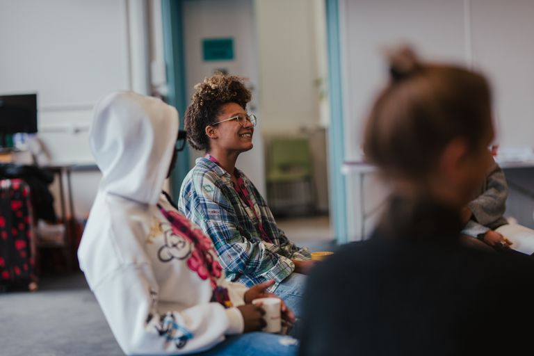 Two women sit next to each other in a circle, one is holding a cup of tea looking at the other woman who is smiling at someone across the room