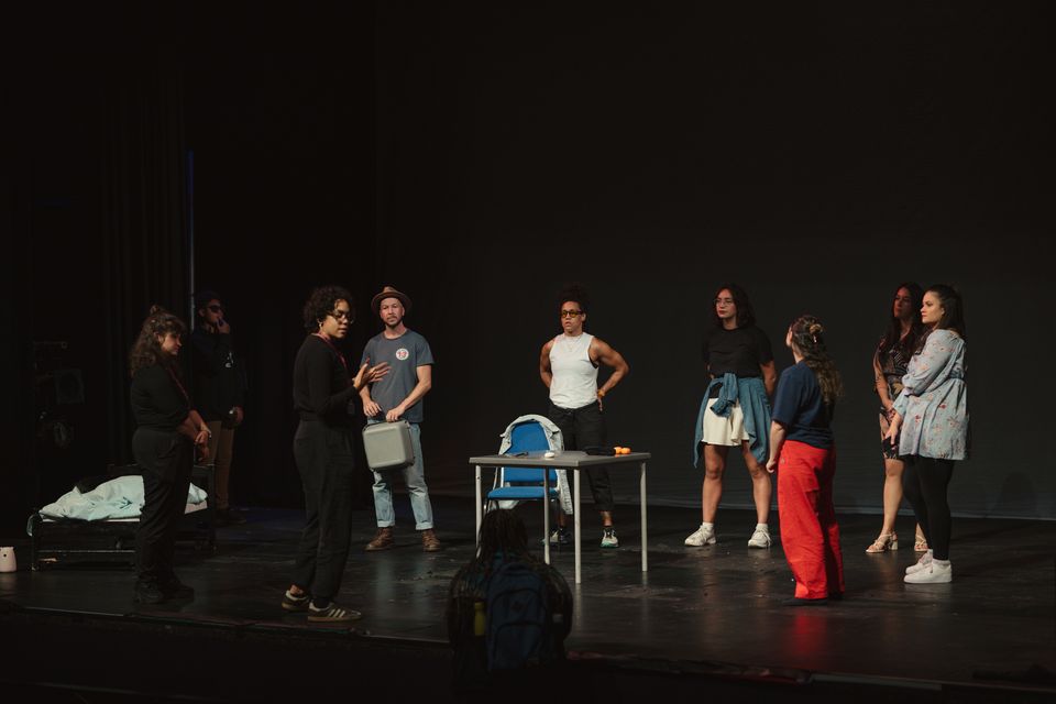 A group of six individuals standing on a stage during a performance. One person gestures as they speak, while the others listen attentively. The stage has minimal props, including a table and a chair. The backdrop is dark, focusing attention on the performers.