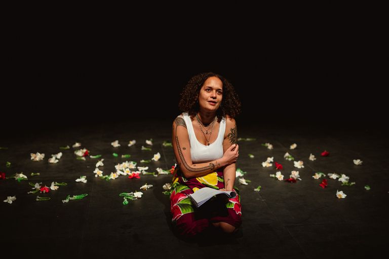 A woman in a white vest top and colourful wrap skirt sits on the stage floor holding a script in her hand, surrounded by flowers