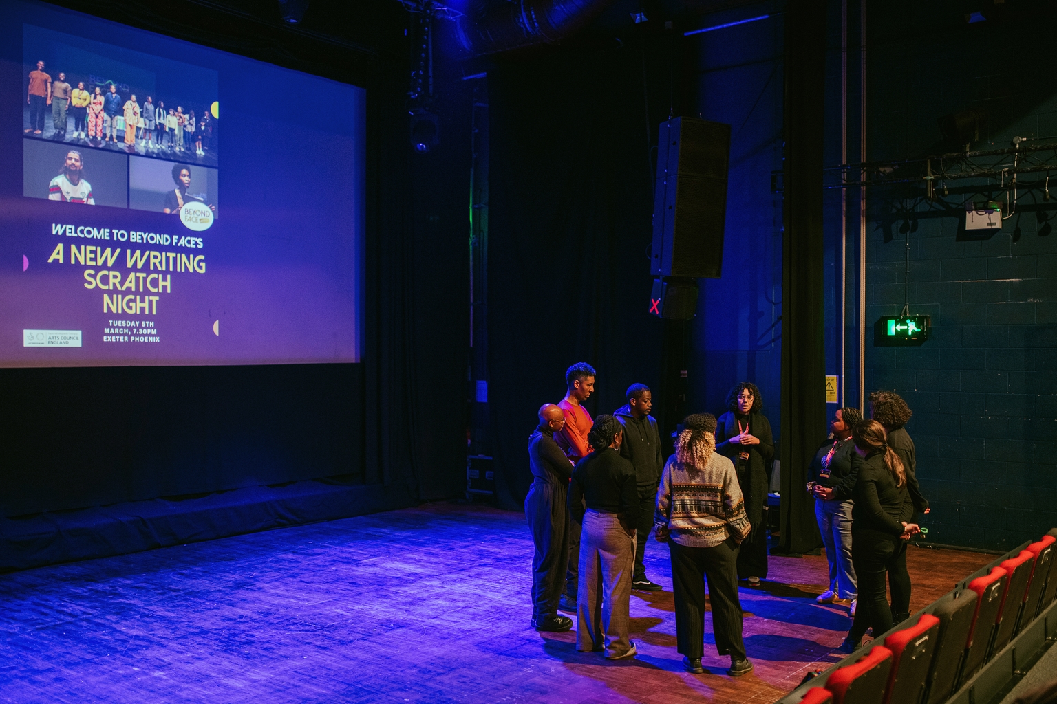 A group of artists debrief in a circle before a show, the words 'New Writing Scratch Night' is projected onto the walls behind them