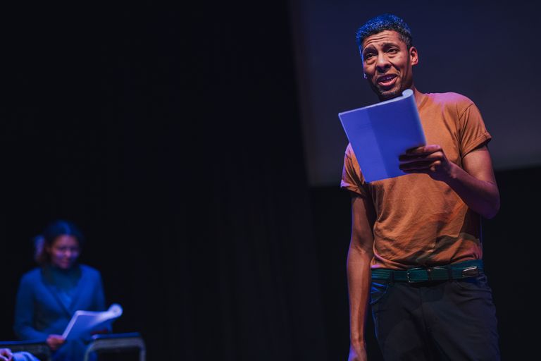 A man with short black hair is on stage holding a script